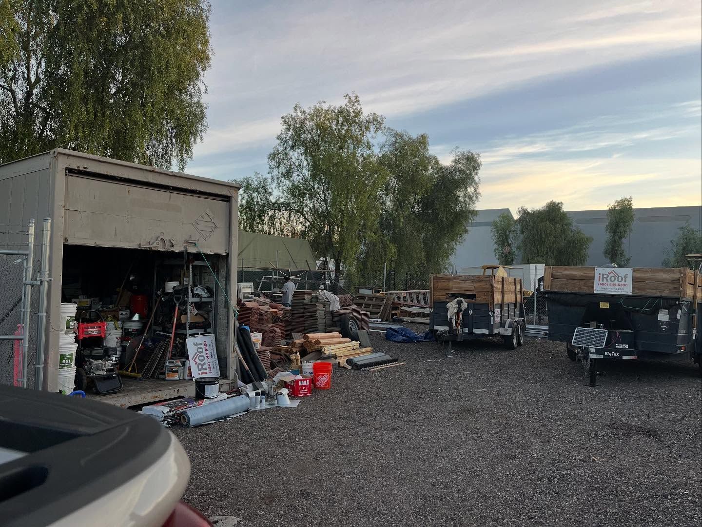 A cluttered outdoor storage area with a partially open shed, piles of materials, and trailers under a cloudy sky.