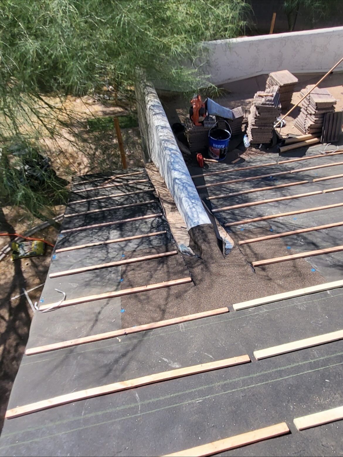 Rooftop construction with black underlayment, wooden slats, and construction materials near a wall and trees.