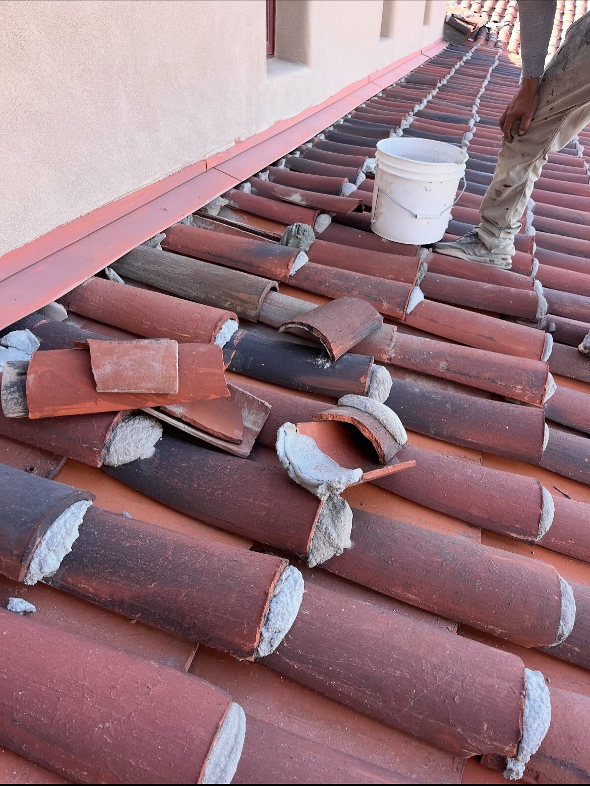Roofer replacing broken terracotta tiles on a red-tiled roof, bucket nearby.