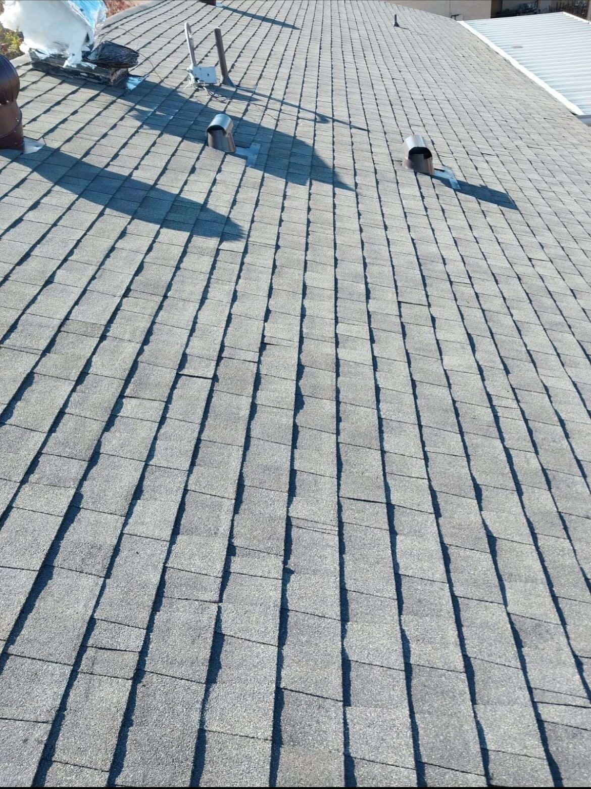 Overhead view of a gray asphalt shingle roof with vents and a satellite dish. Sunny.