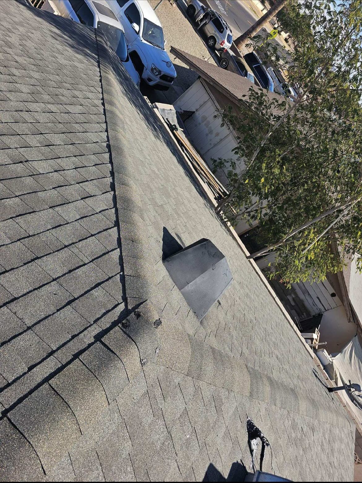 Dark asphalt shingle roof with a vent, viewed from above, surrounded by buildings and cars.