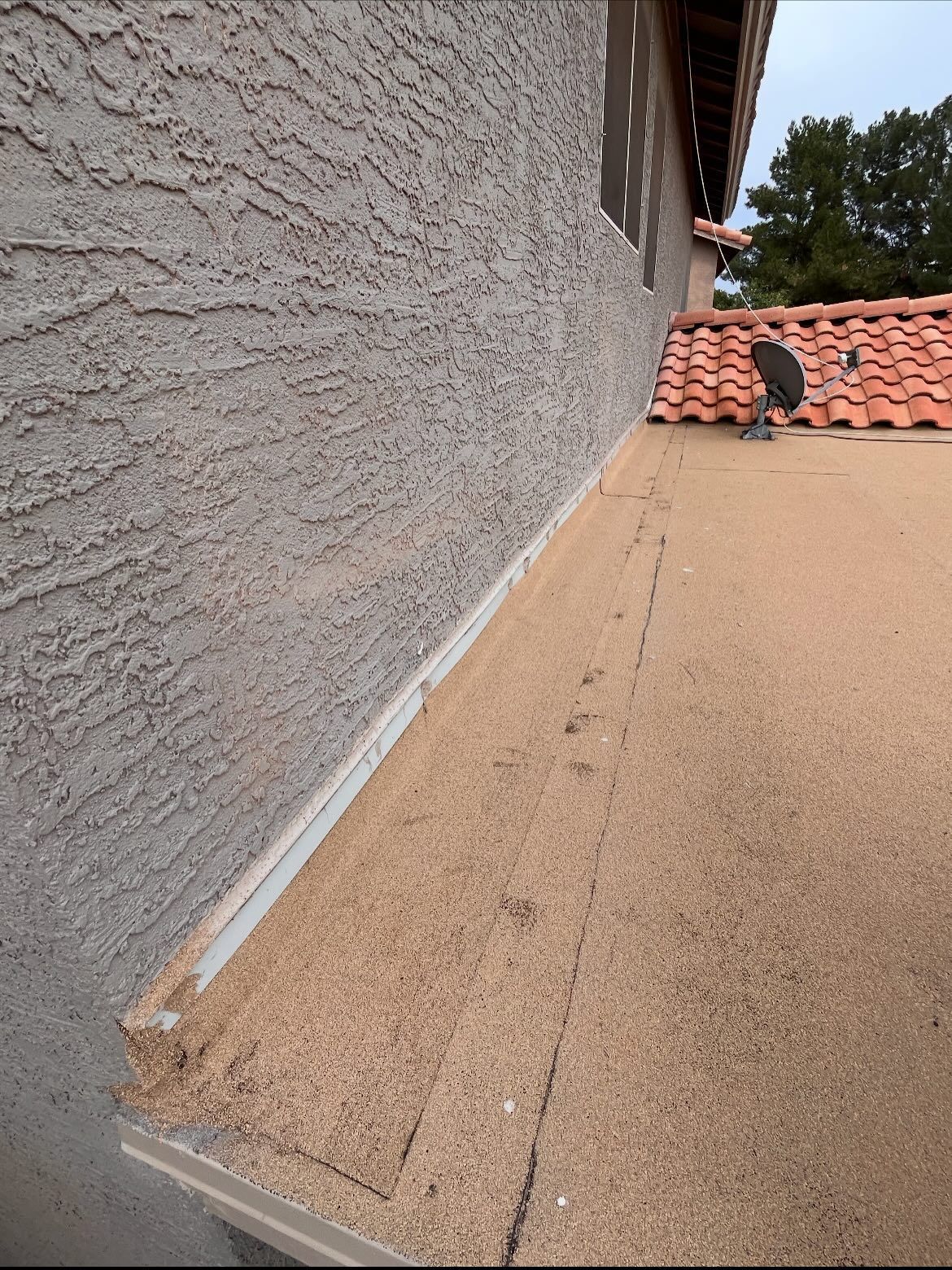 Exterior view of a stucco wall and flat roof. Terra-cotta roof tiles in the background. Light brown hues.