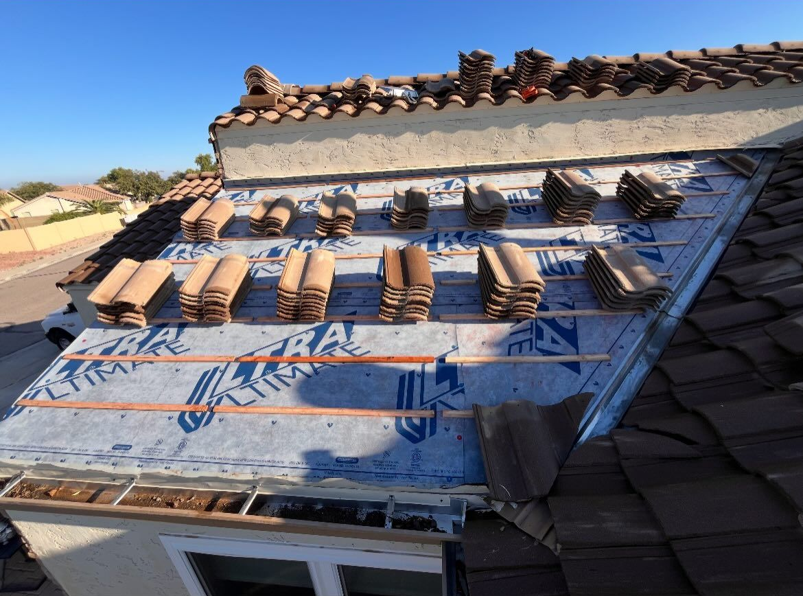 Roof in progress; brown tiles stacked on blue underlayment and wooden battens. Bright sky.