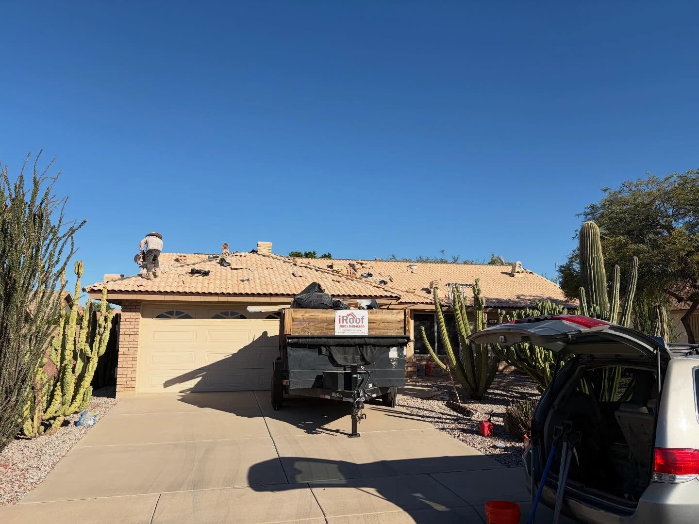 A house with roof damage, a trailer in the driveway, and a clear blue sky.