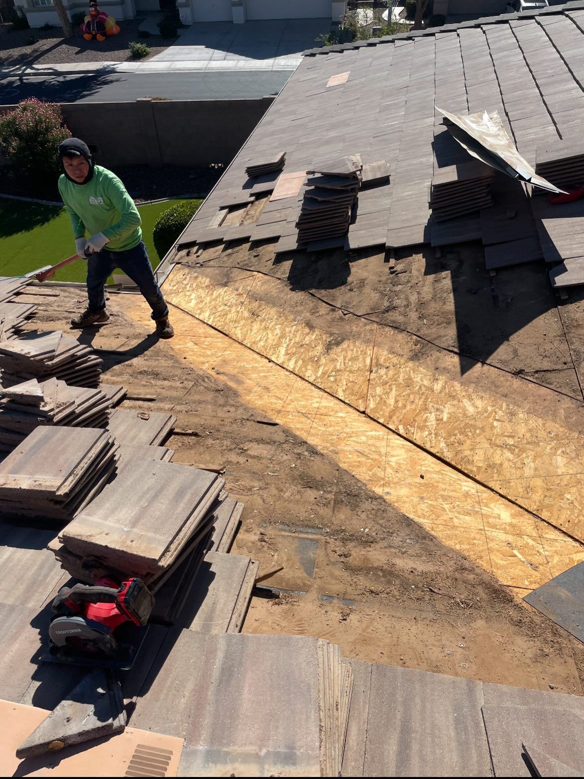 Man on a rooftop replacing tiles; section of roof missing tiles, revealing wood.