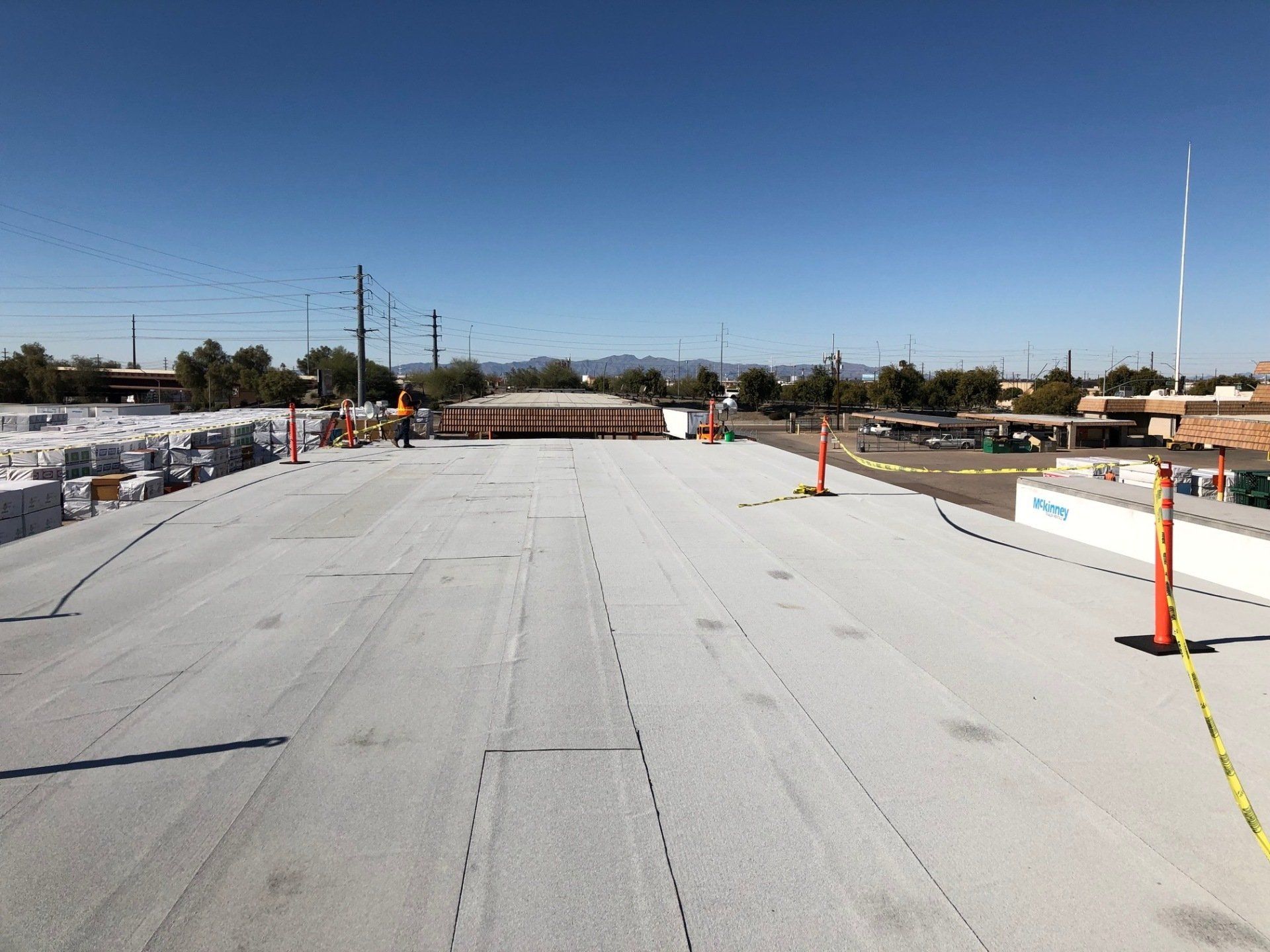 Flat roof construction site with white material, cones, and workers under a blue sky.