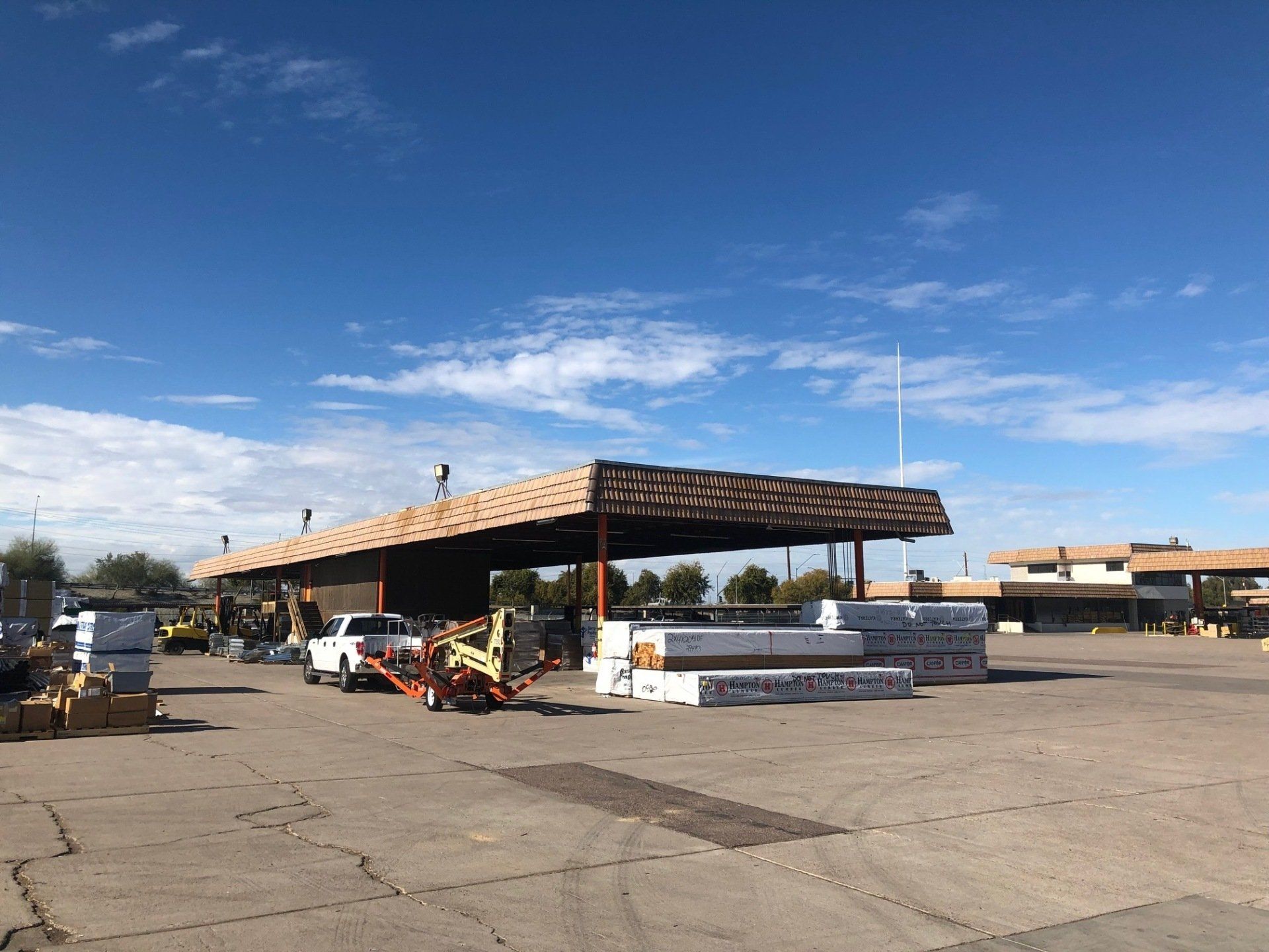Lumber yard with stacks of wood under a covered area, vehicles, and a clear blue sky.
