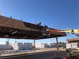Workers on a lift repairing a building roof on a sunny day. Lumber stacks are in the background.