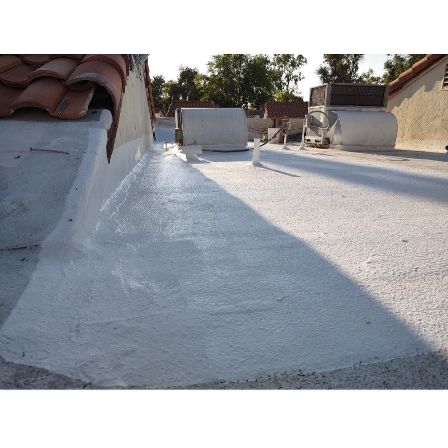 White coated flat roof with a stucco wall and red tile roof, visible air conditioning units in background.