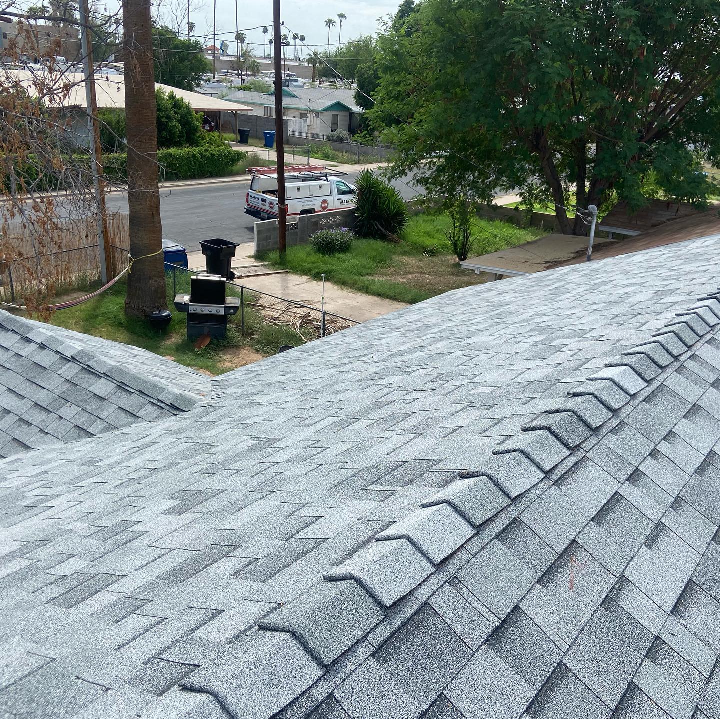 Gray shingled roof with a view of a street, sidewalk, trees, and parked utility vehicles.