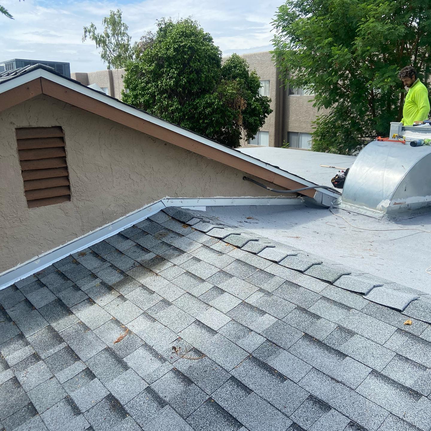 Roofer on a roof with gray asphalt shingles and a flat, silver metal section, sunny day.