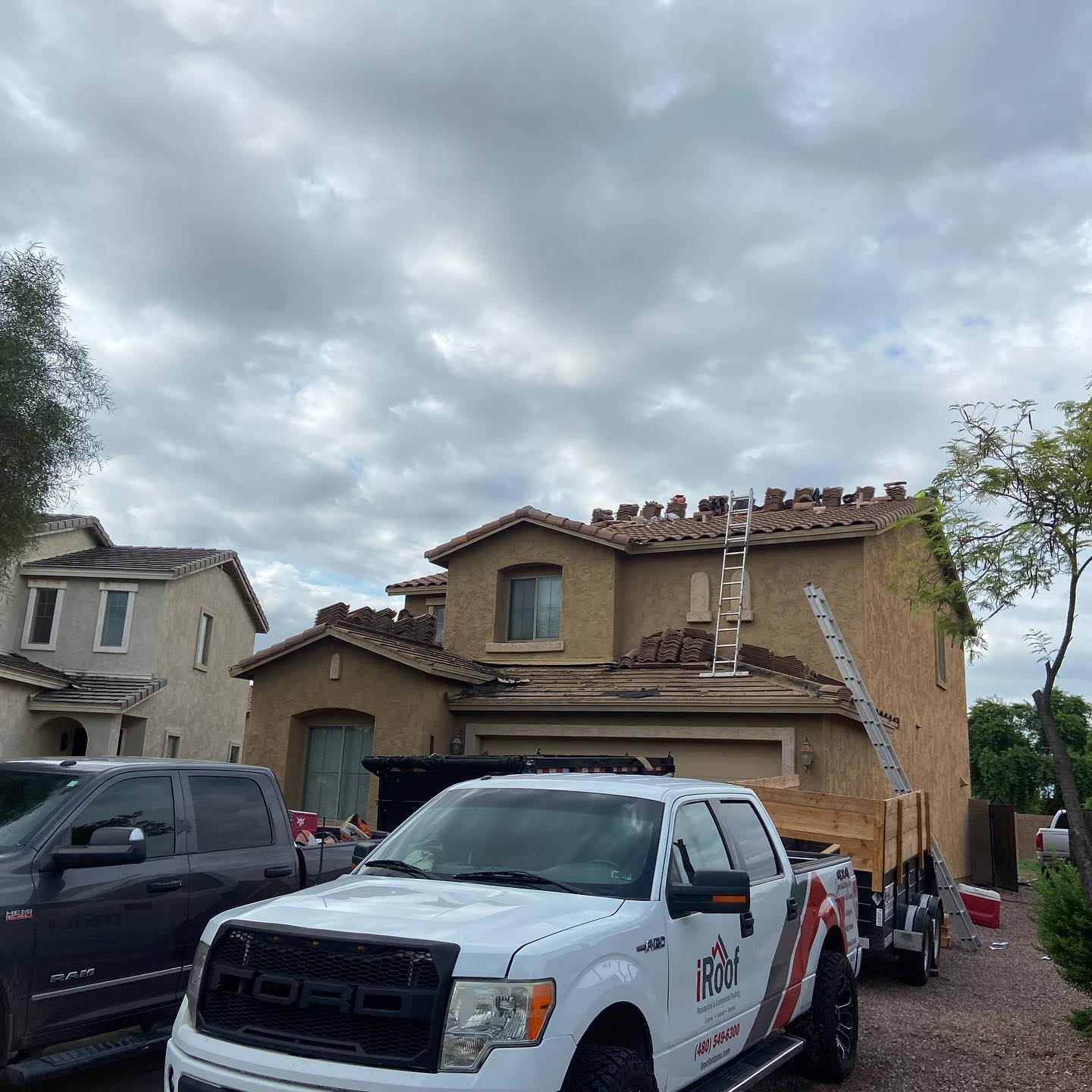 A two-story house with roof replacement in progress, surrounded by trucks and equipment under a cloudy sky.