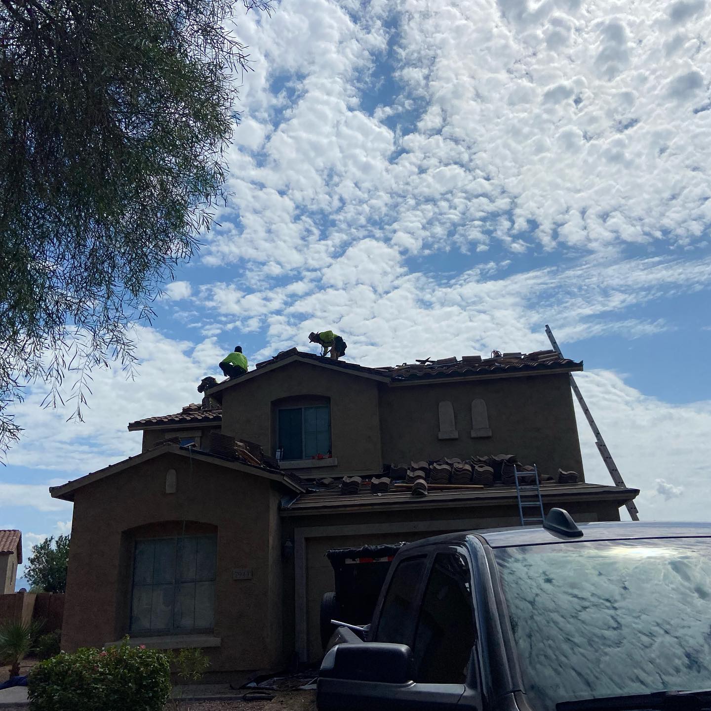 Two workers on a house roof under a cloudy blue sky, next to a black vehicle.