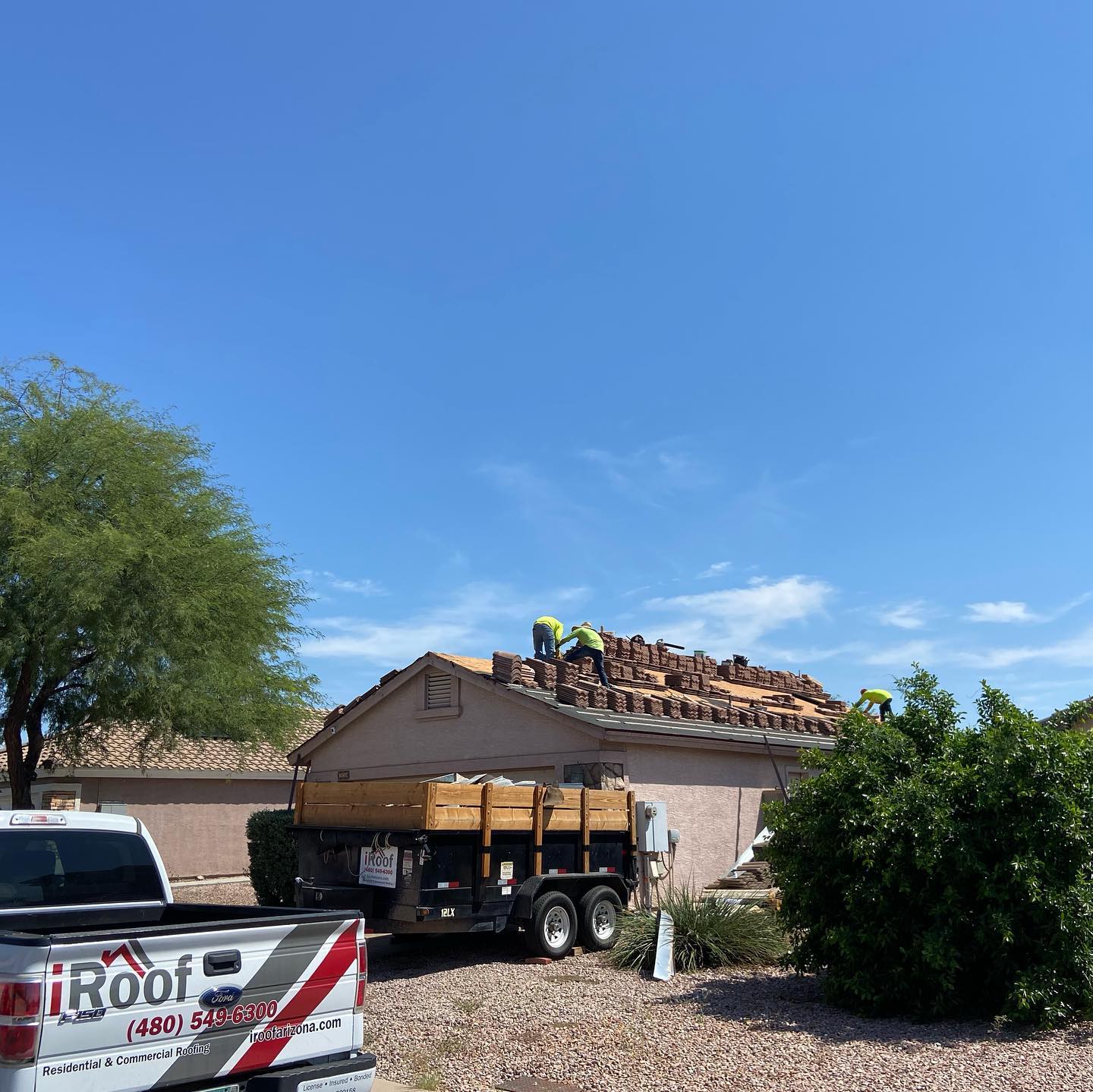 Roofers replacing tiles on a house under a clear blue sky. A truck and trailer are present.