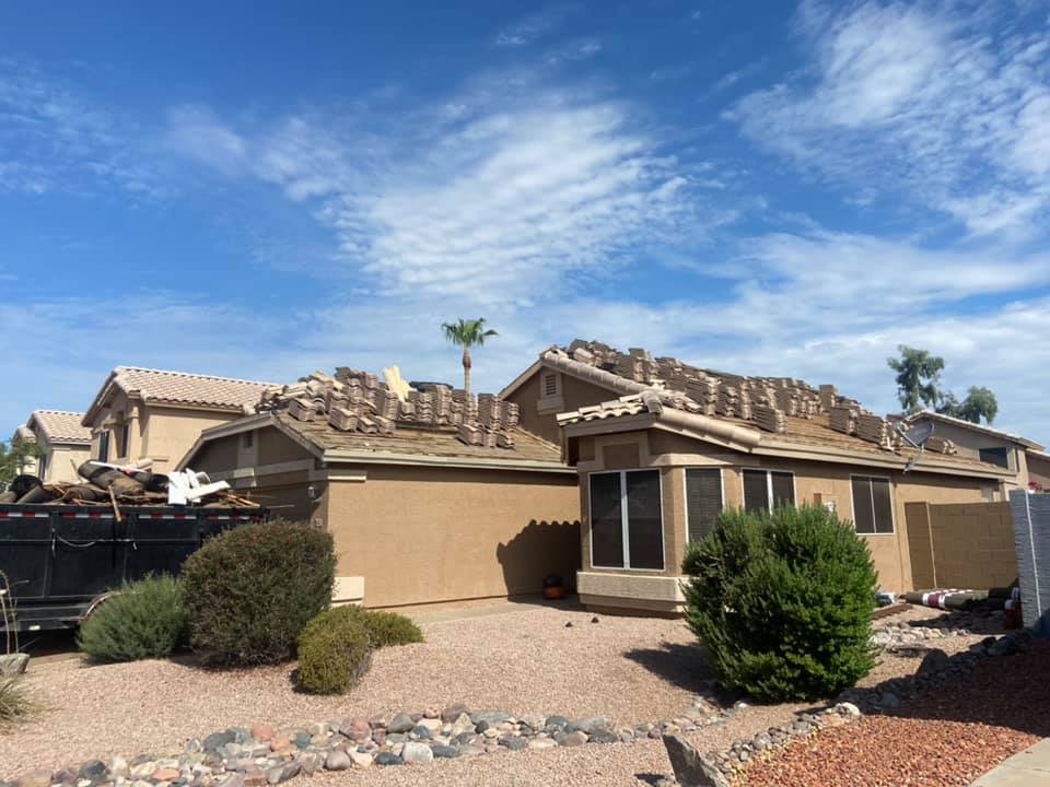 House with roof partially removed, debris visible. Blue sky, sunny day. Brown house, tan gravel yard.