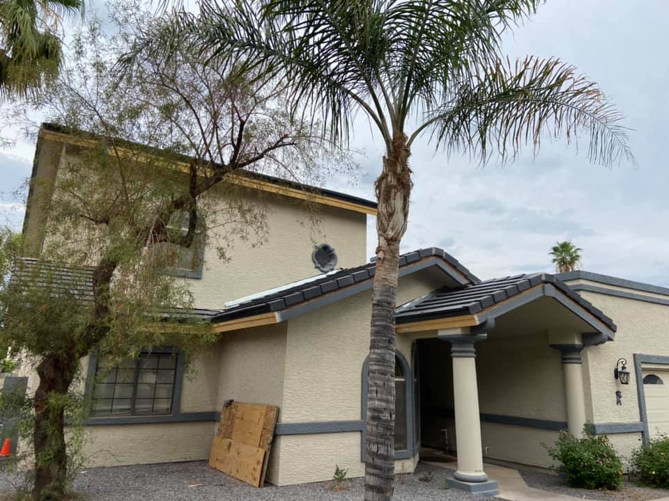 Two-story house with stucco exterior, dark roof, and palm trees. Cloudy sky. Wood panel propped against the wall.