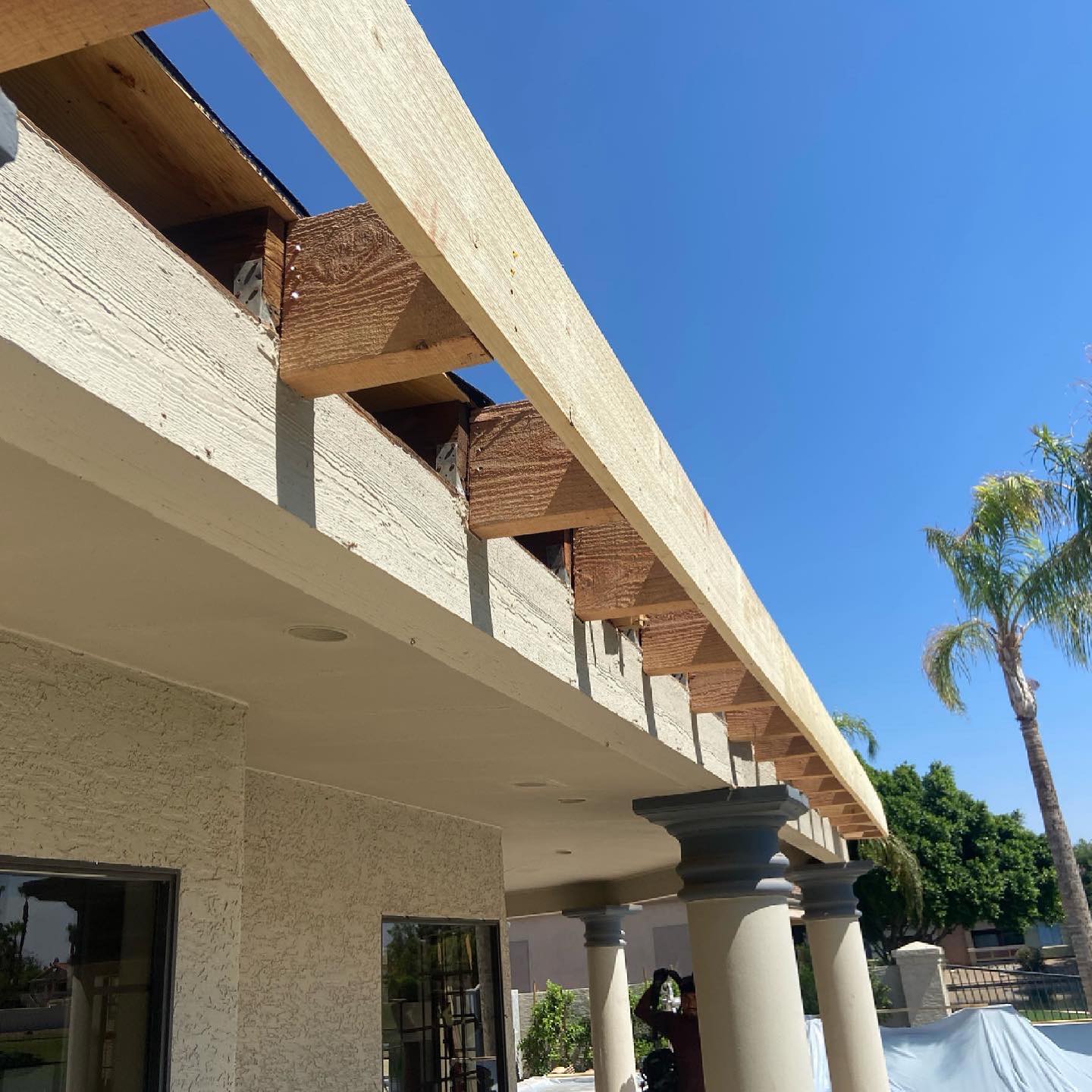 View of a patio with a partial roof under construction, featuring wooden beams and stucco columns against a clear blue sky.