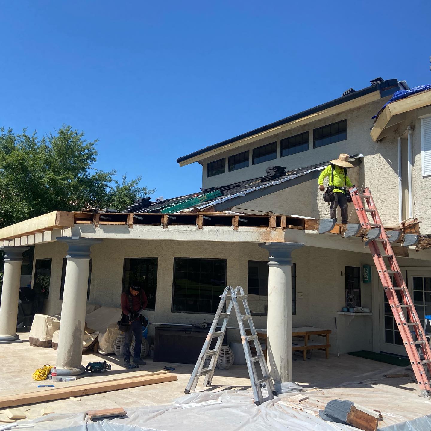 Construction workers on a house with a damaged roof. One is on a ladder, others below. Blue sky.