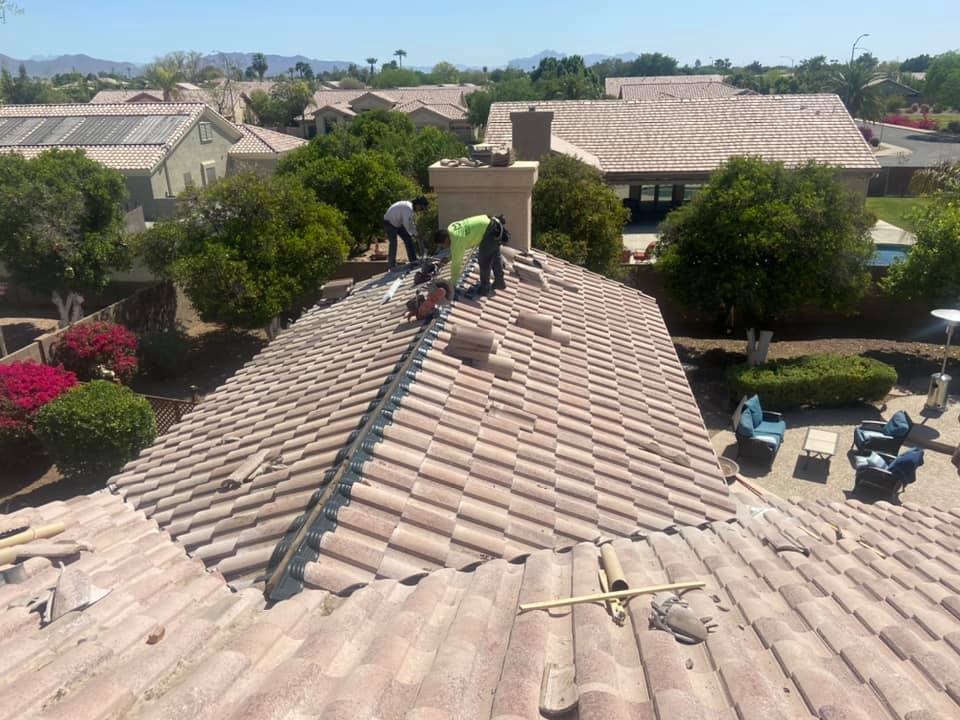 Workers on a clay-tiled roof repairing the structure near a chimney; sunny day, residential neighborhood.