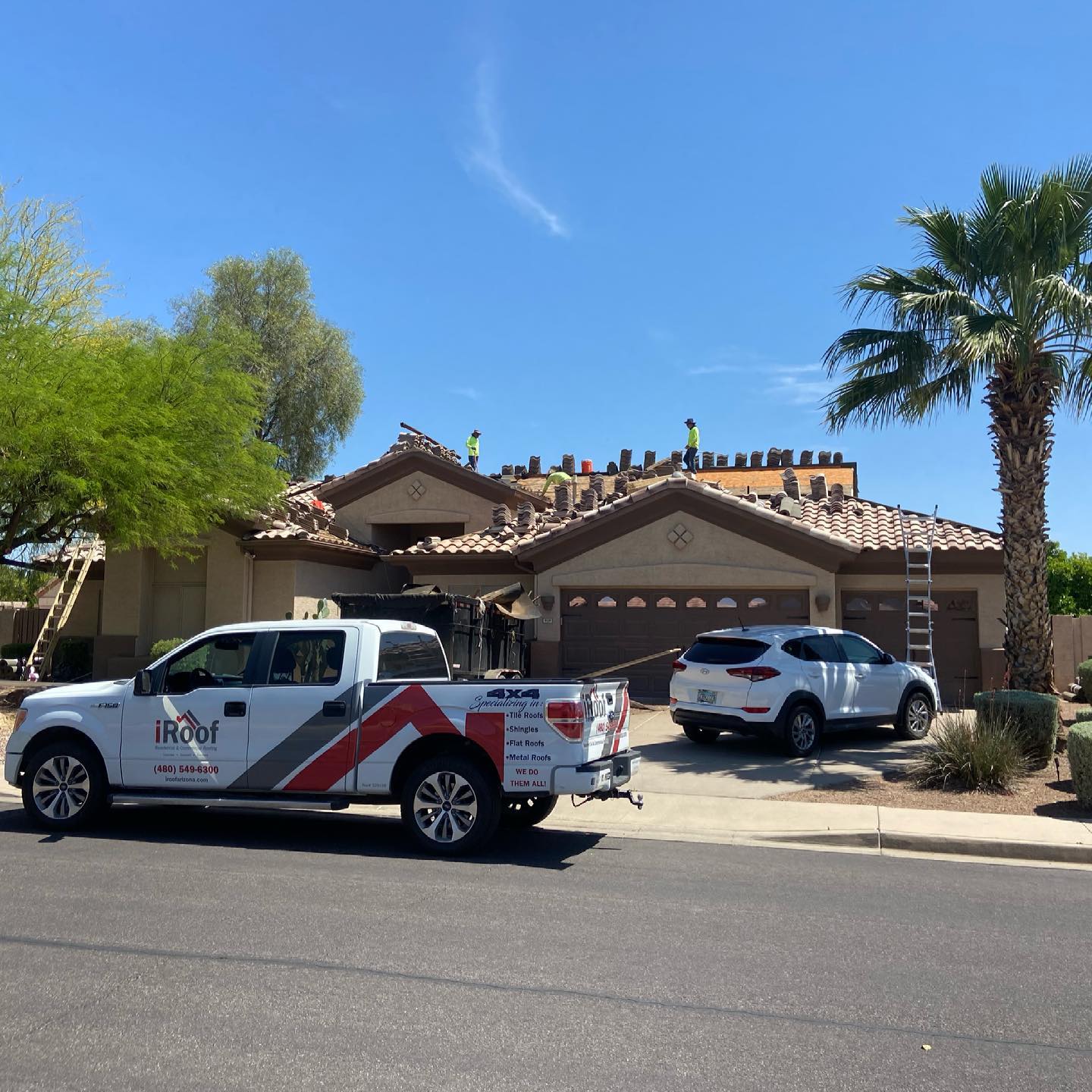 Roofers on a house with a white truck, SUV, palm tree, and clear blue sky.