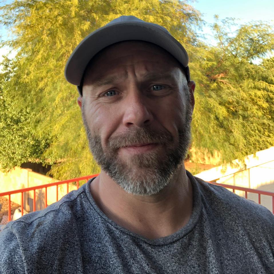 Man with a gray hat and beard, wearing a gray shirt, smiles outdoors with a tree in the background.