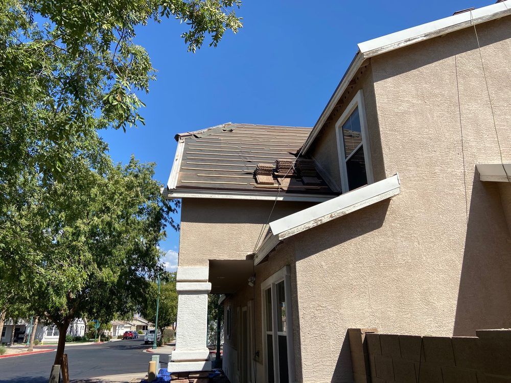 Damaged roof on a tan stucco house with a white trim. Bright blue sky overhead.