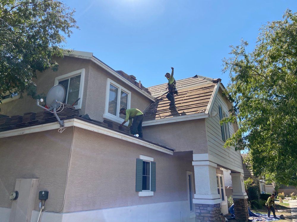 Workers on a roof replacing shingles on a two-story house under a clear blue sky.