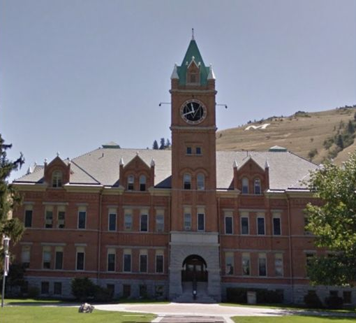 A brick building with a clock tower under a clear sky. A hill is visible in the background.