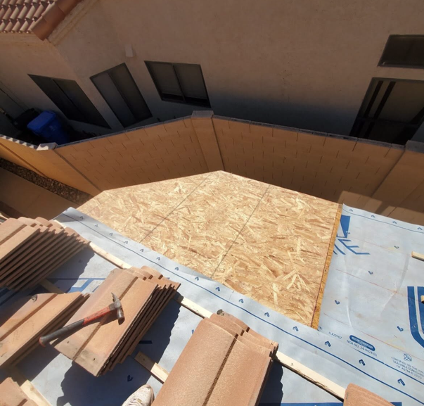 Rooftop construction: OSB sheathing on a gable end, with tiles and hammer nearby. Blue underlayment visible.