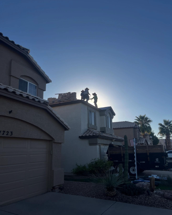 Two people working on a roof, silhouetted against a bright sky, near a residential area.