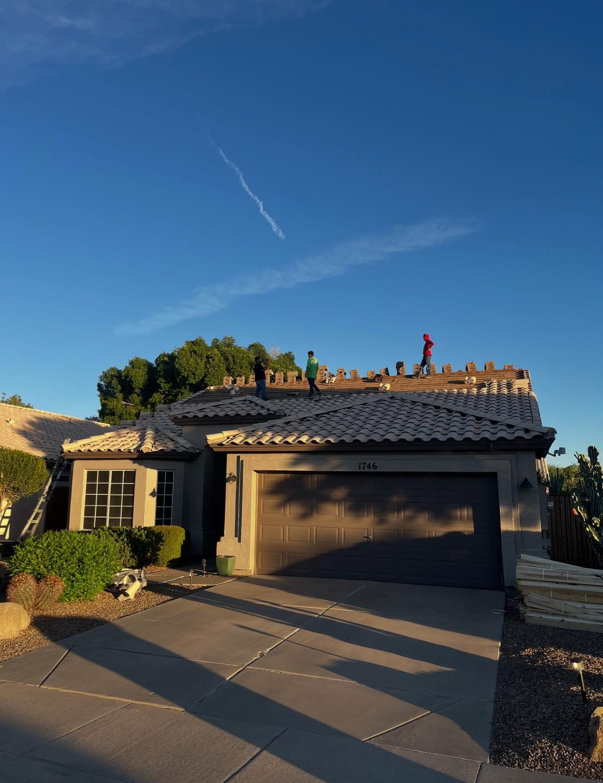 Workers on a roof with tan tiles, under a clear blue sky, replacing tiles on a house with a brown garage door.