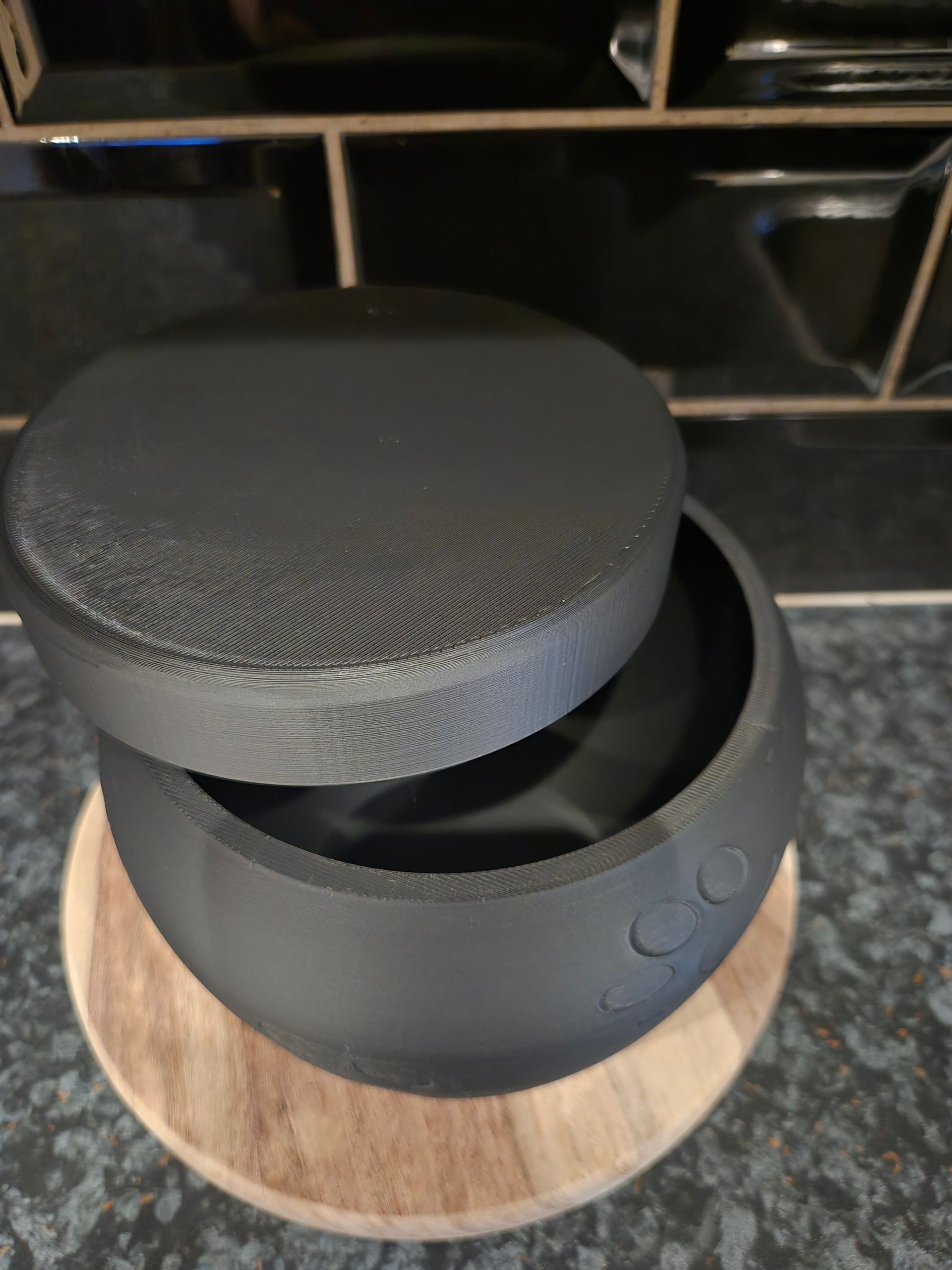 Een matzwarte cilindervormige luidspreker met een licht geopende bovenklep, rustend op een ronde houten voet op een aanrecht.