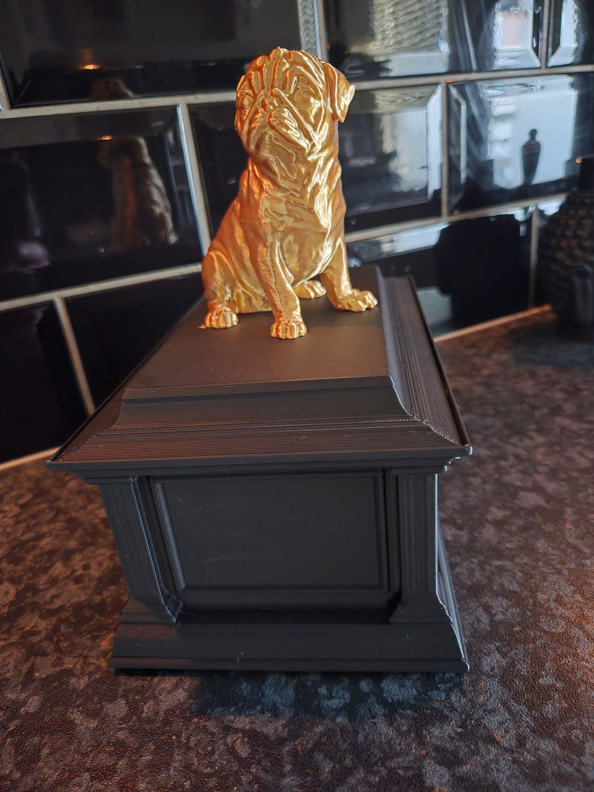 Een klein goudkleurig mopshondje zit bovenop een donkergrijze rechthoekige crematieurn op een donkere ondergrond.