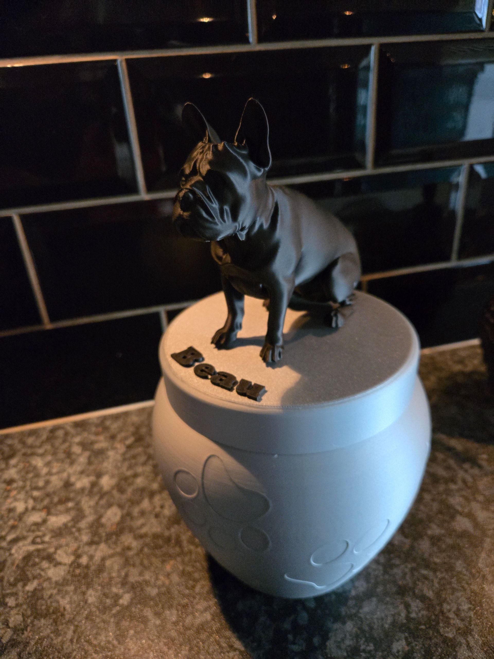 Een matwitte urn met een gegraveerd patroon op de voet en een donkermetalen beeldje van een Franse bulldog bovenop het deksel.