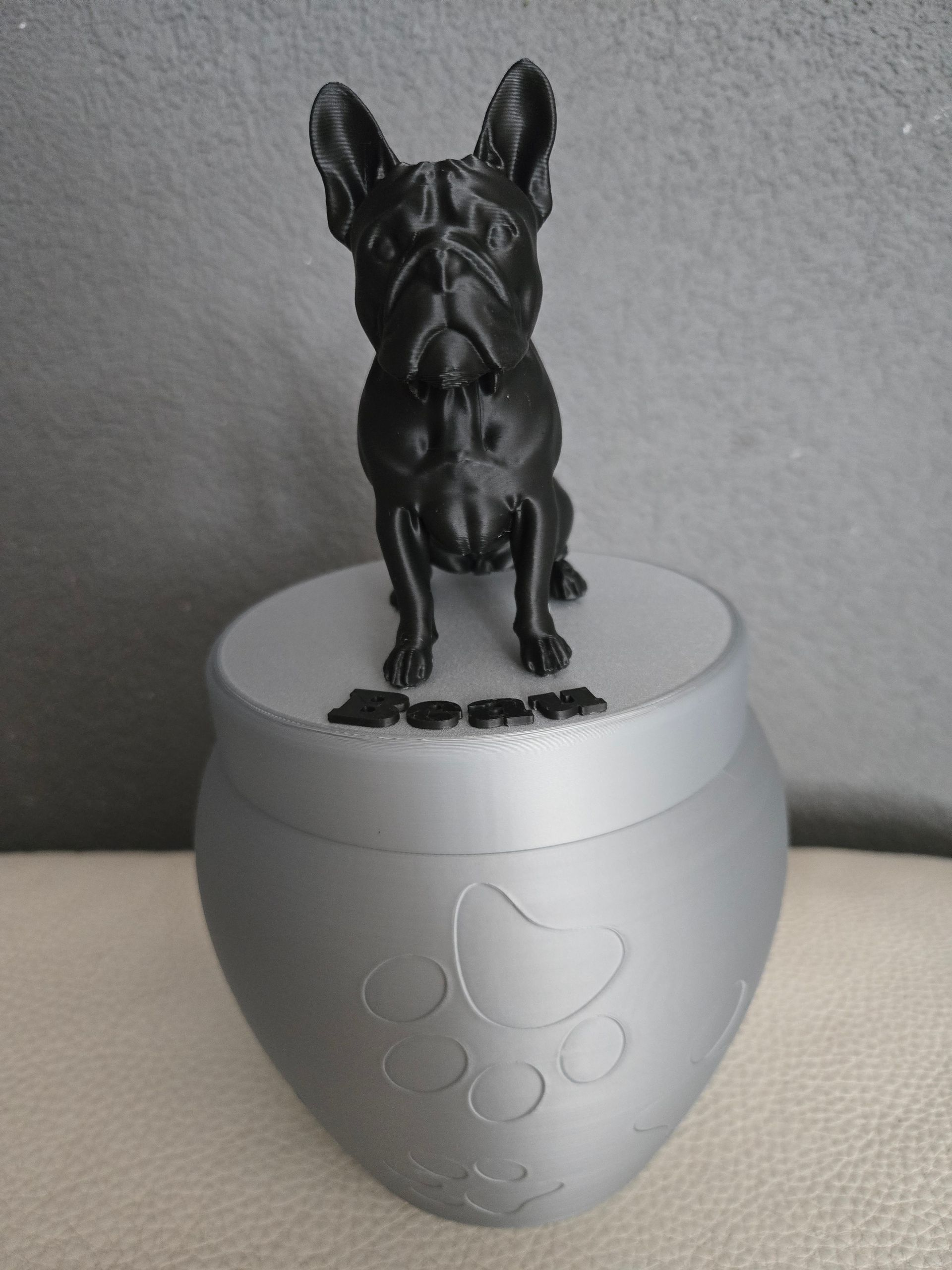 Een zilveren dierenurn met een reliëf van een pootafdruk, bekroond met een zwart 3D-geprint beeldje van een Franse bulldog en de naam
