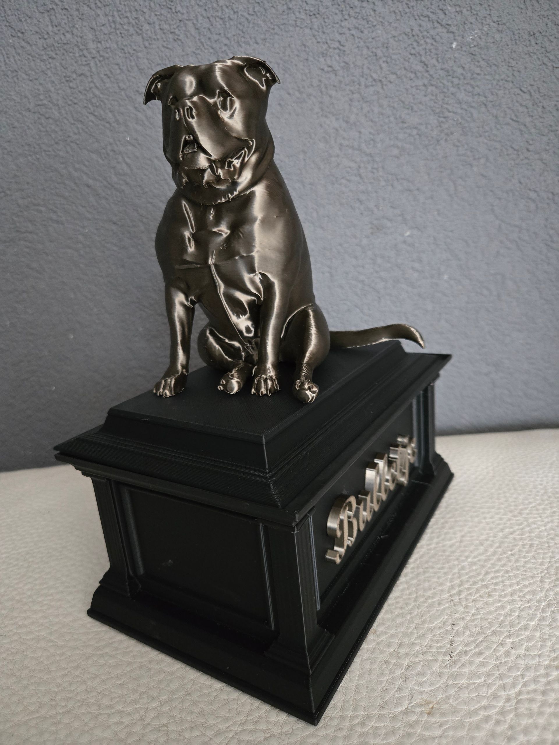 Een bronskleurig hondenbeeldje dat op een zwart rechthoekig voetstuk staat met de naam 