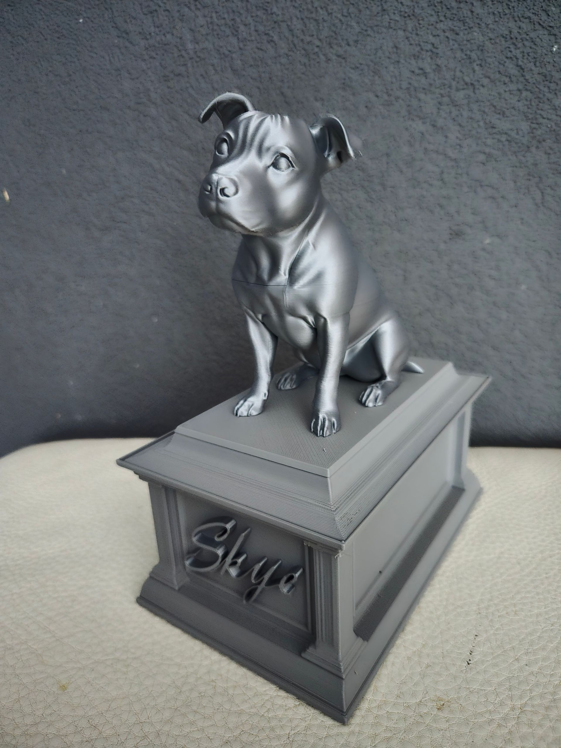 Een donkergrijze, 3D-geprinte gedenkurn met een zittend hondenbeeldje erop, waarop de naam 