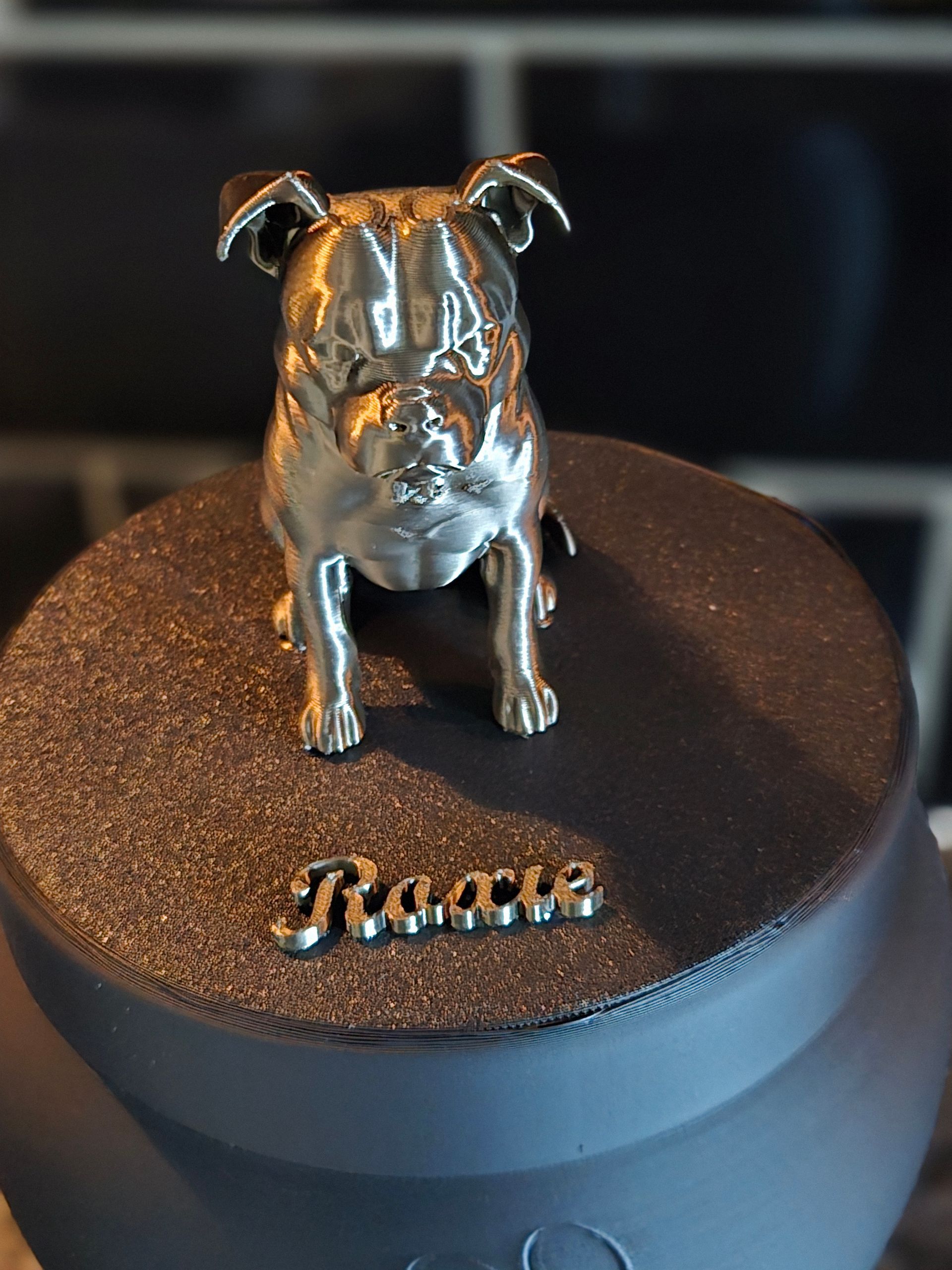 Een metalen beeldje van een bulldog staat op een donker, gestructureerd potdeksel met het opschrift 