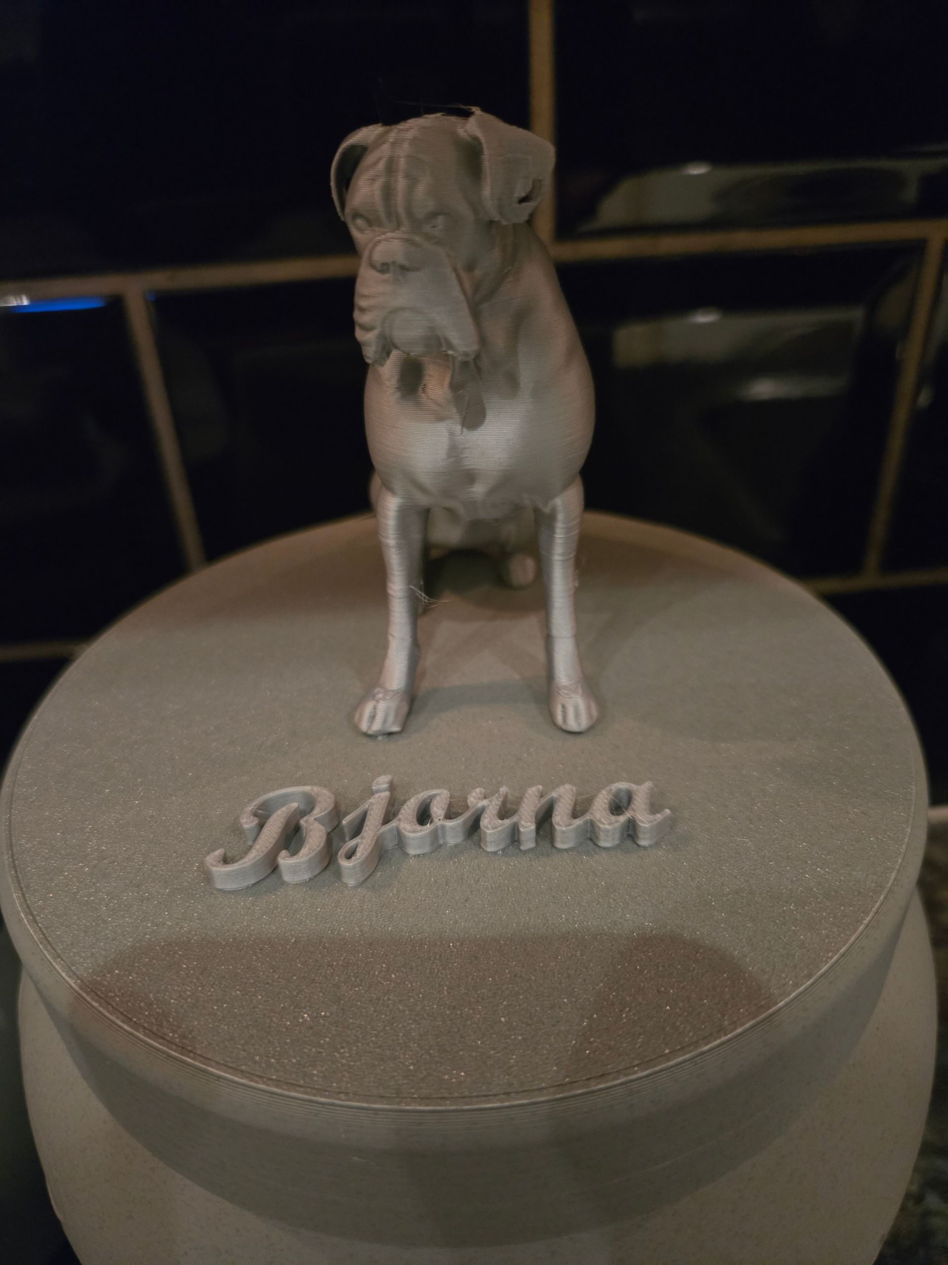 Een lichtgekleurd, 3D-geprint beeldje van een Boxer staat op een ronde urnvormige voet met de naam 