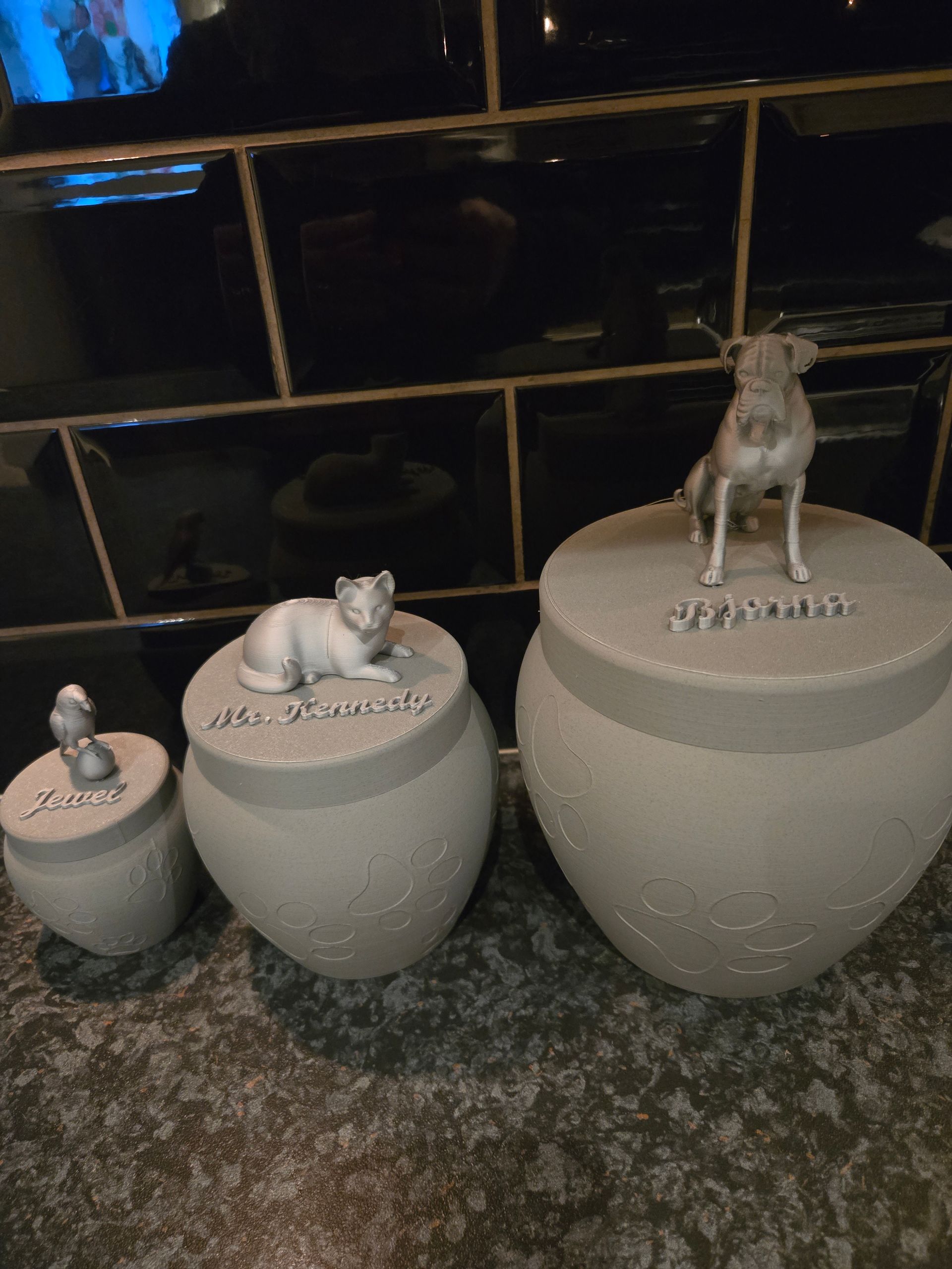 Drie gestructureerde, steenkleurige urnen met deksels van verschillende afmetingen, elk bekroond met een klein dierenbeeldje, op een donkere ondergrond.