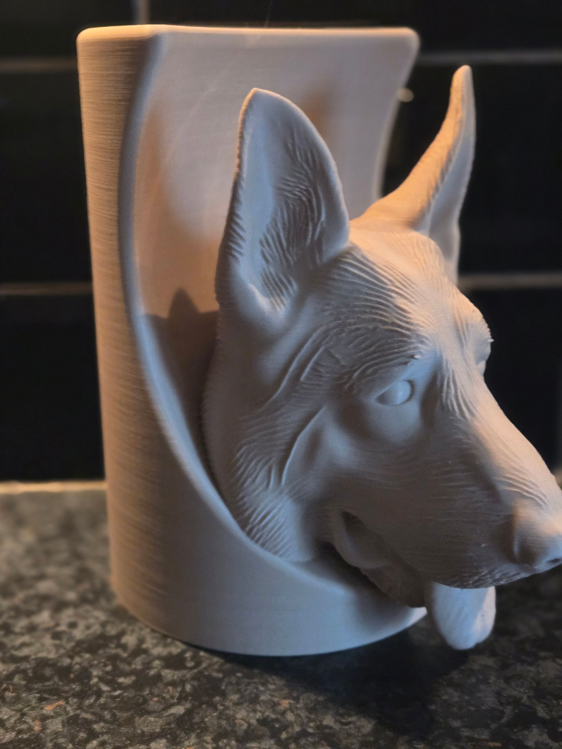 Een 3D-geprinte, witte, keramische kop van een Duitse herder die uit een gebogen cilindrische voet op een granieten ondergrond steekt.