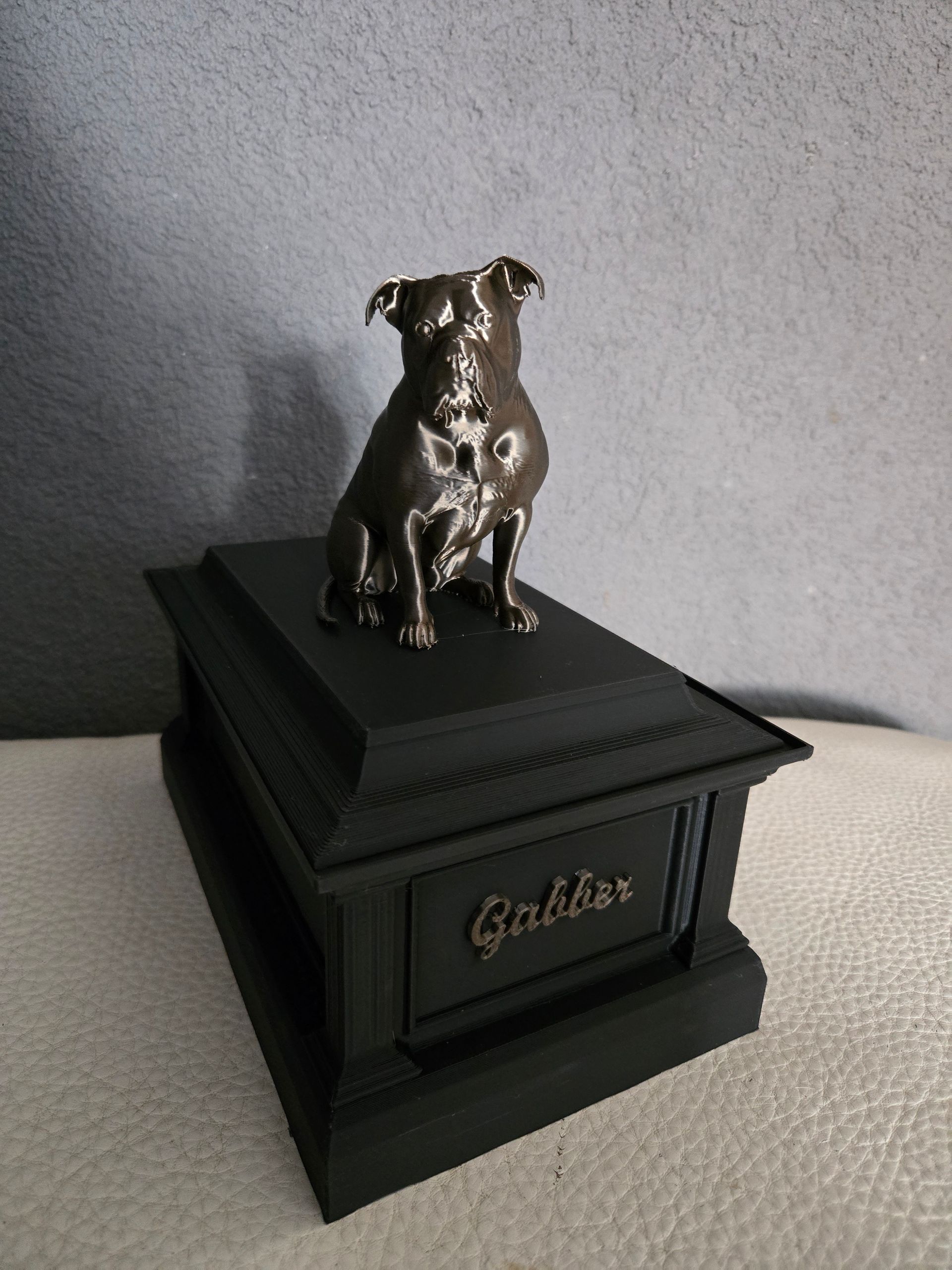 Een kleine, matzwarte dierenurn met een bronskleurig hondenbeeldje bovenop en de inscriptie 