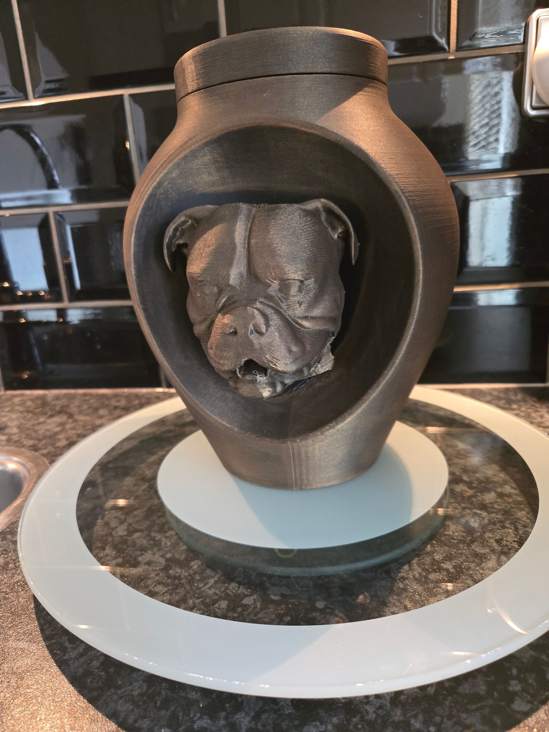 Een donkere, metallic, 3D-geprinte urn met een gebeeldhouwde bulldogkop in het midden, rustend op een witte ronde voet.