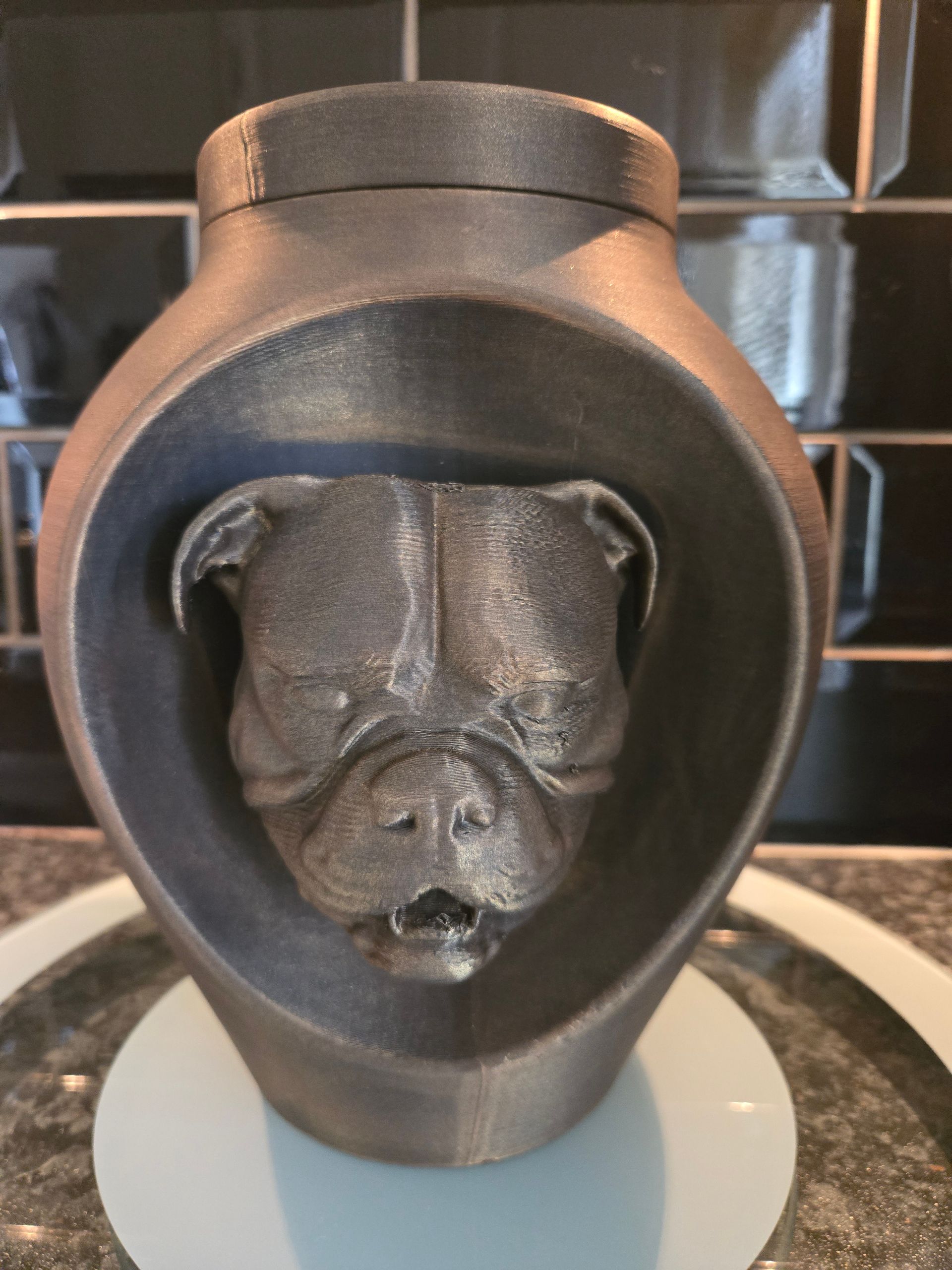 Een 3D-geprinte, houtskoolkleurige urn met een gebeeldhouwd gezicht van een Franse bulldog in het midden van een verzonken ovale omlijsting.