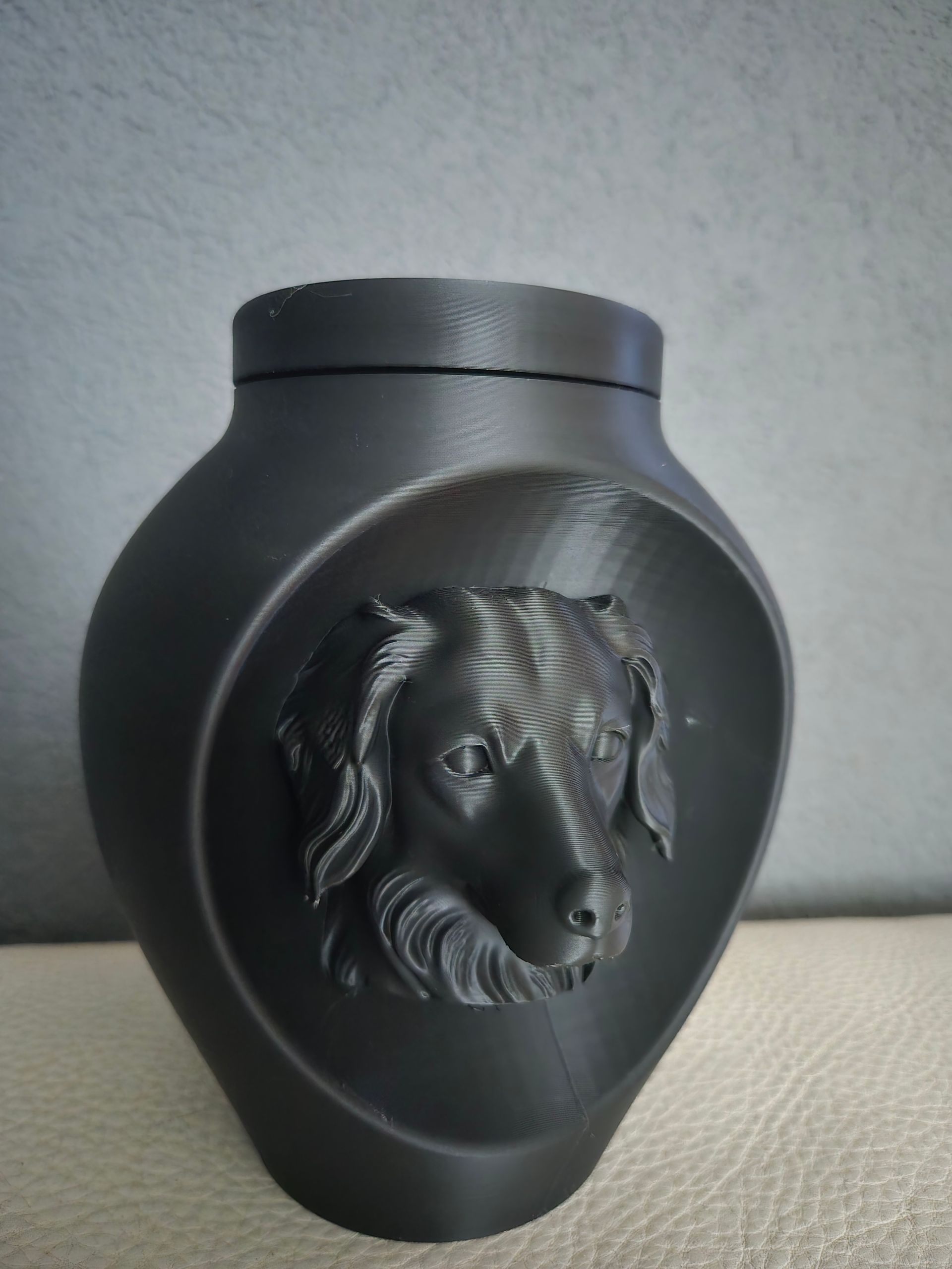 Een matzwarte, 3D-geprinte urn met een reliëf van een hondenkop op de voorkant, tegen een effen grijze achtergrond.