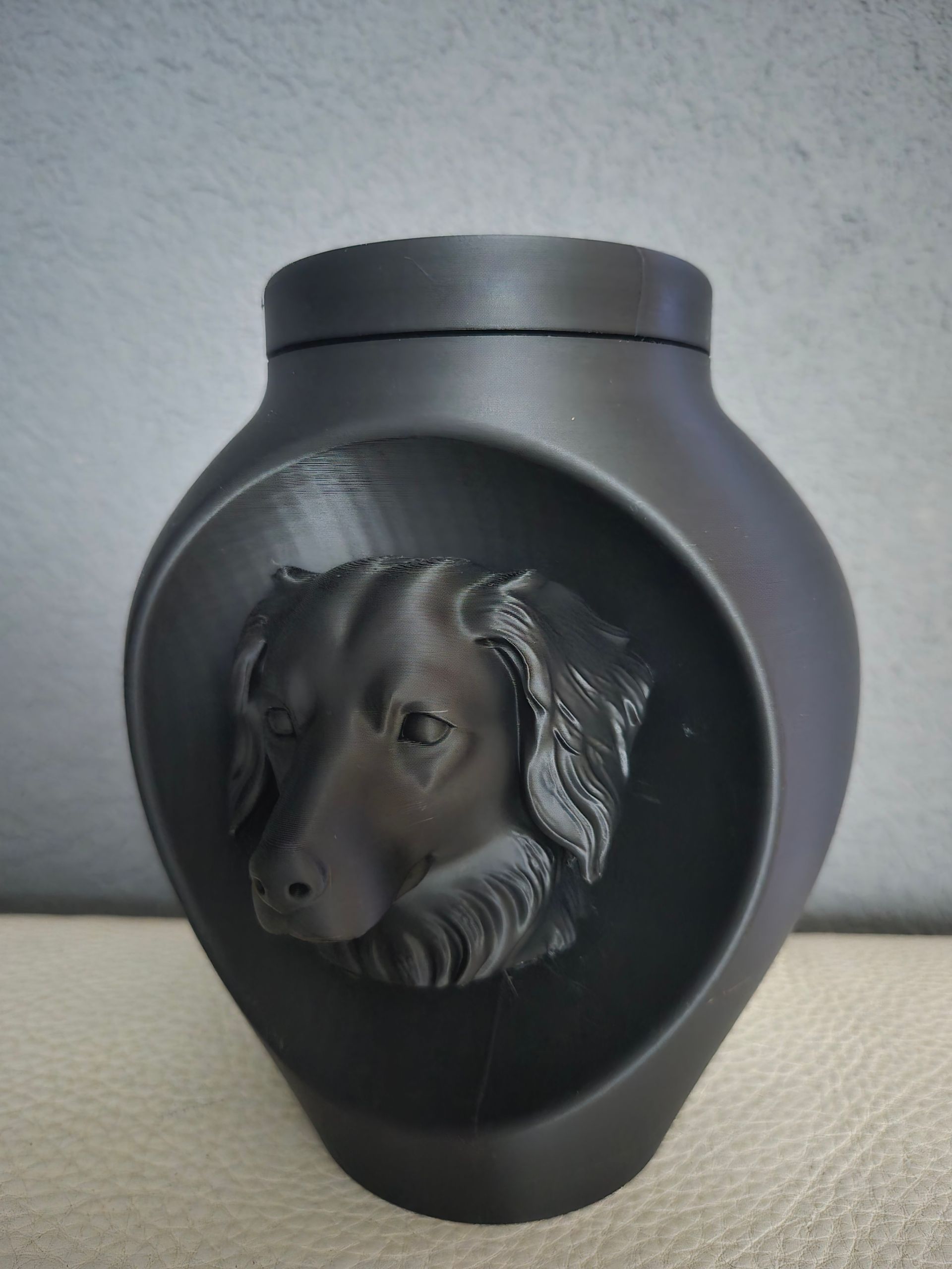 Een zwarte, matte dierenurn met een reliëf van een hondenkop aan de voorkant, tegen een lichtgrijze achtergrond.