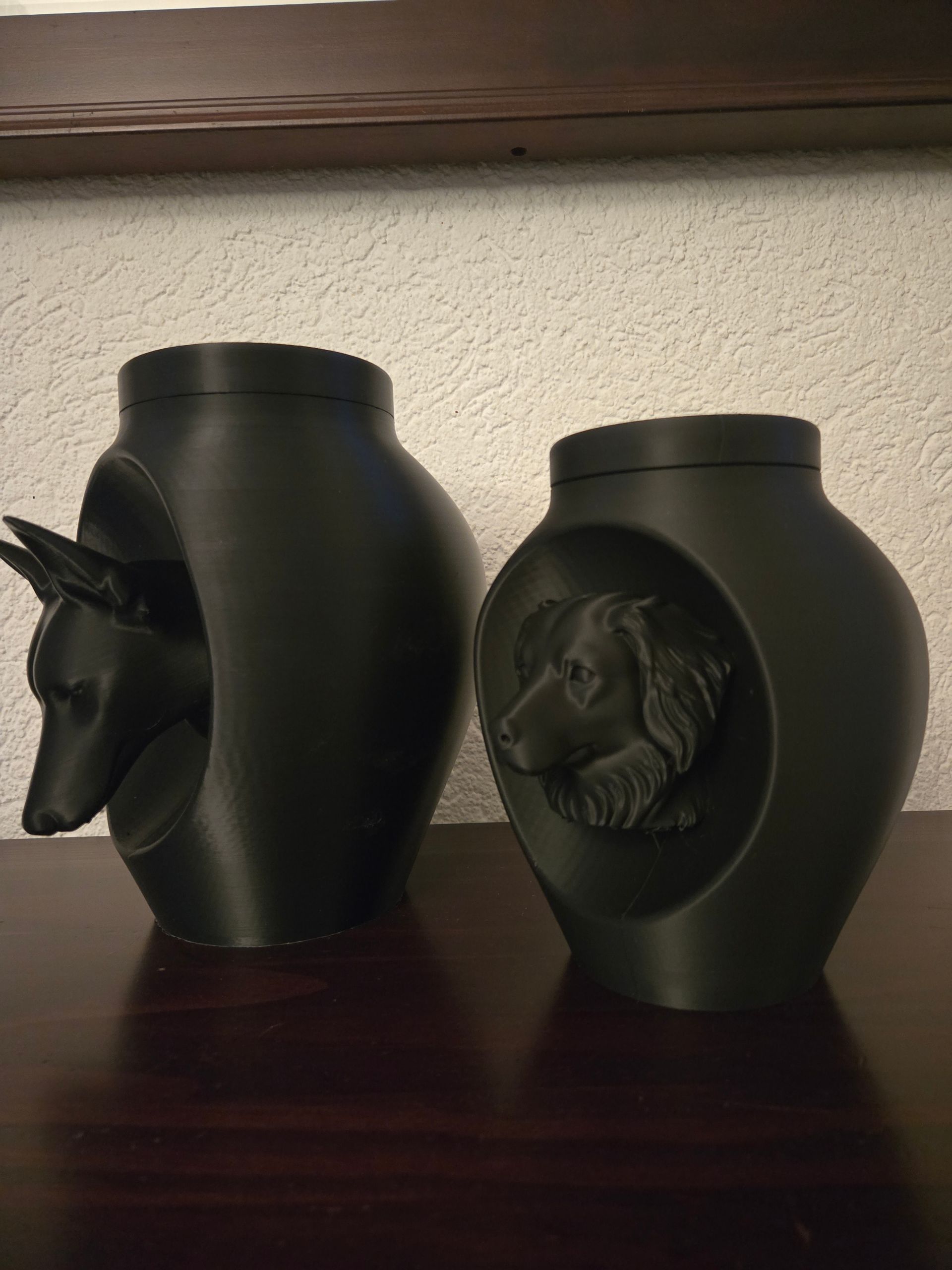 Twee matzwarte, vaasvormige dierenurnen, elk met een reliëfsculptuur van een ander hondenras, staan ​​op een tafel.