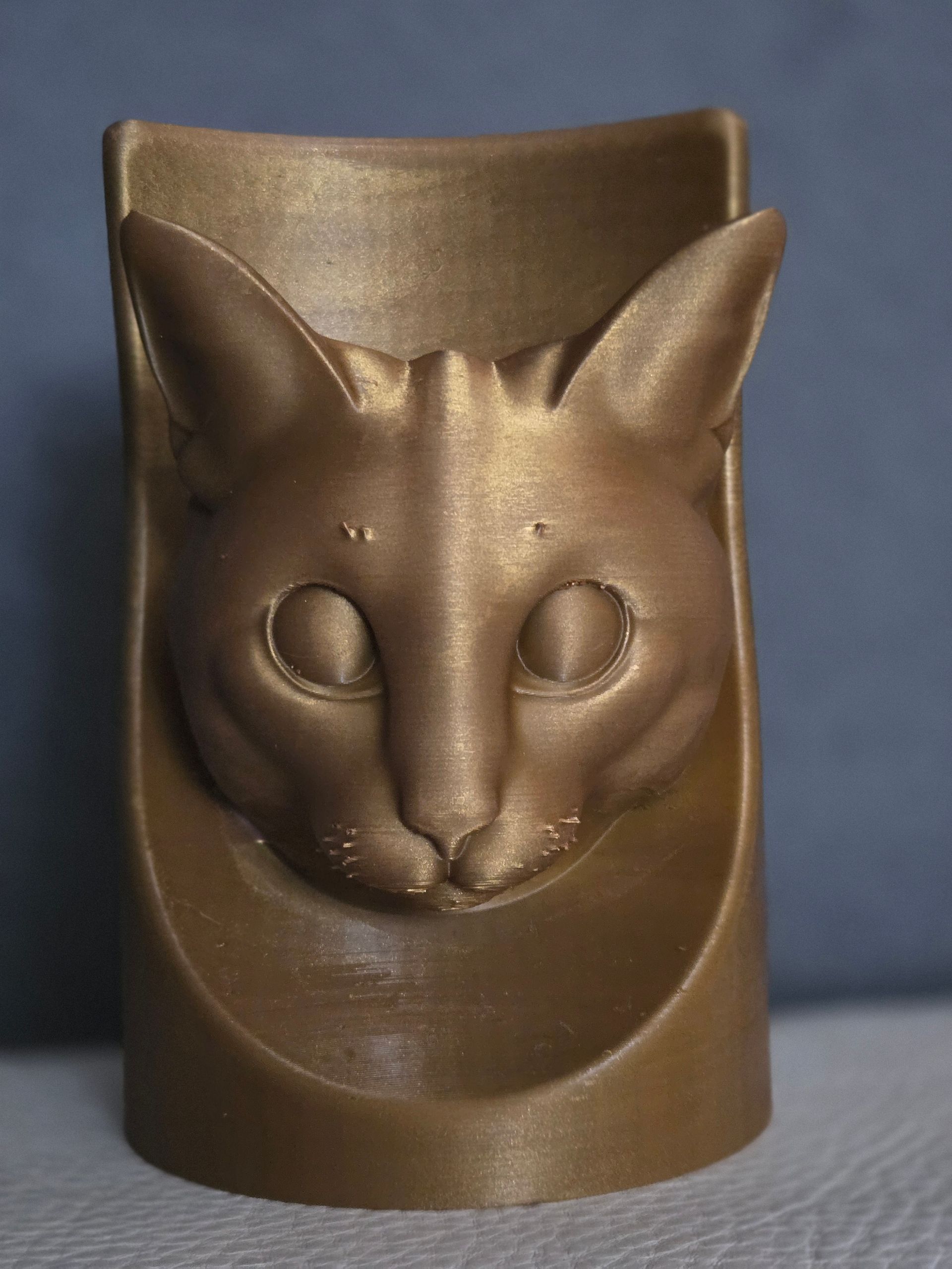 Een bronskleurige, 3D-geprinte cilindervormige container met een gebeeldhouwd kattengezicht op de voorkant.