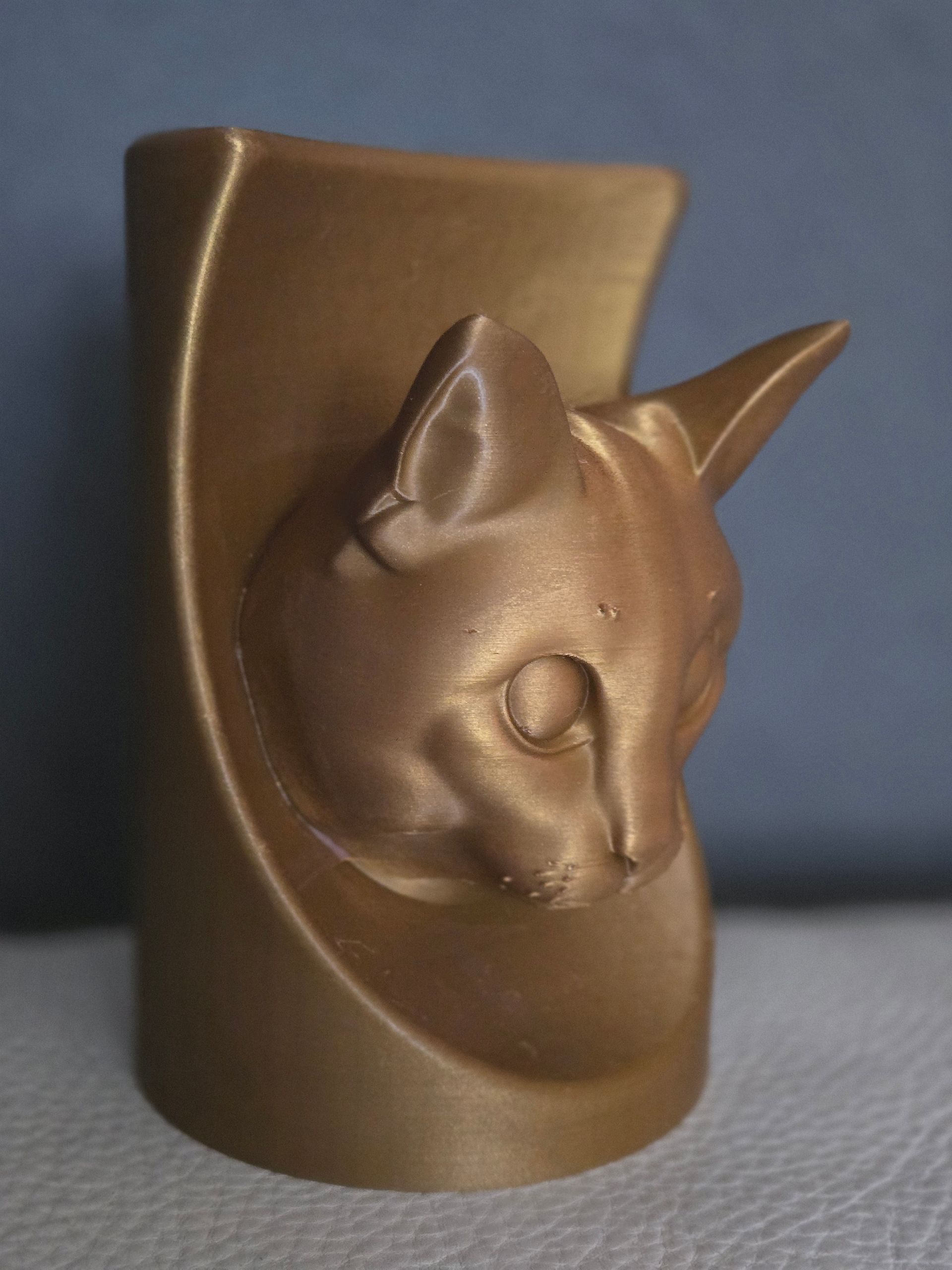 Een cilindrische vaas in metallic bronskleur, 3D-geprint, met een gebeeldhouwde kattenkop die aan de voorkant uitsteekt.