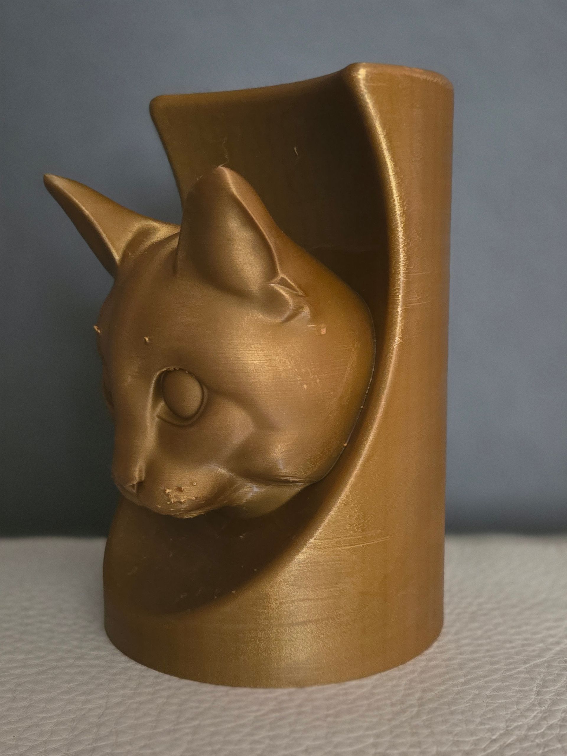 Een 3D-geprinte sculptuur van een kattenkop in metallic goud, die uit een gebogen, cilindrische standaard tevoorschijn komt.